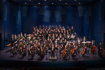 A stage filled with an orchestra and people playing violins, cellos, a choir, drums, and a conductor  