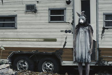 A still from a scary movie of an old RV and a girl standing in front of it with a bunny mask 