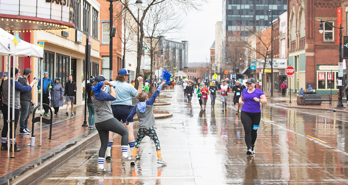 a woman running down Barstow Street at the Eau Claire Marathon while the crowd cheers her on by clapping and shaking pom-poms in the air 