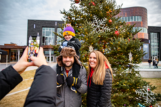 A family of 3 posing in front of a statue of Santa and the holiday tree in Haymarket Plaza  