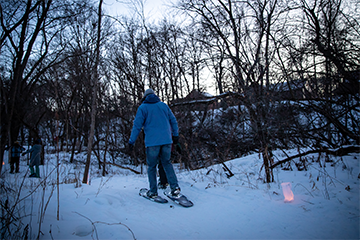 Man walking into the woods on snowshoes through a candlelit, snow covered trail at dusk 