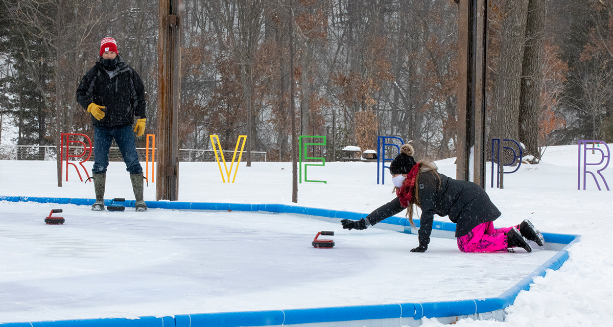 Woman wearing snowpants and jacket on her knees on the ice playing Crokicurl in River Prairie while Man on her team watches from the side of the ice 