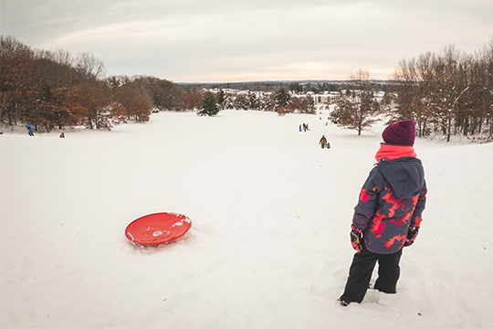 Young girl standing at the top of a snowy hill with snowpants and jacket with her sled, looking at the bottom of the hill she's about to sled down 