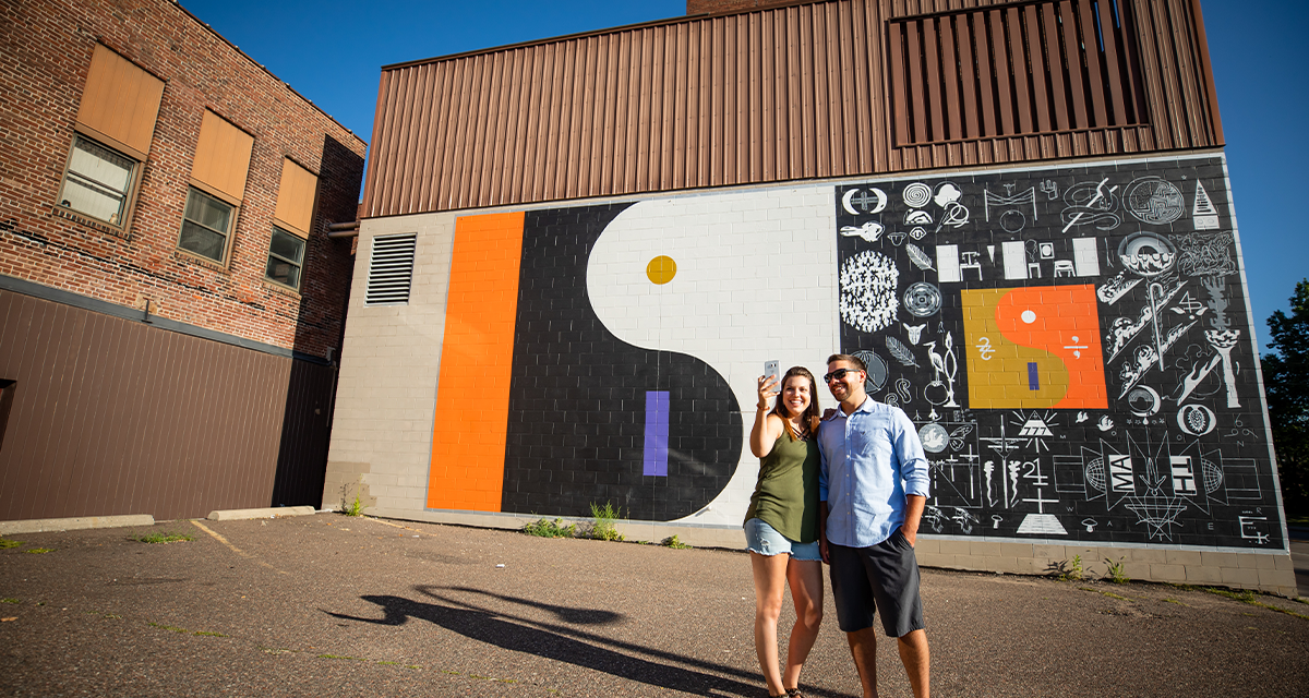 A couple taking a selfie in front of the Bon Iver 22 A Million mural in downtown Eau Claire 