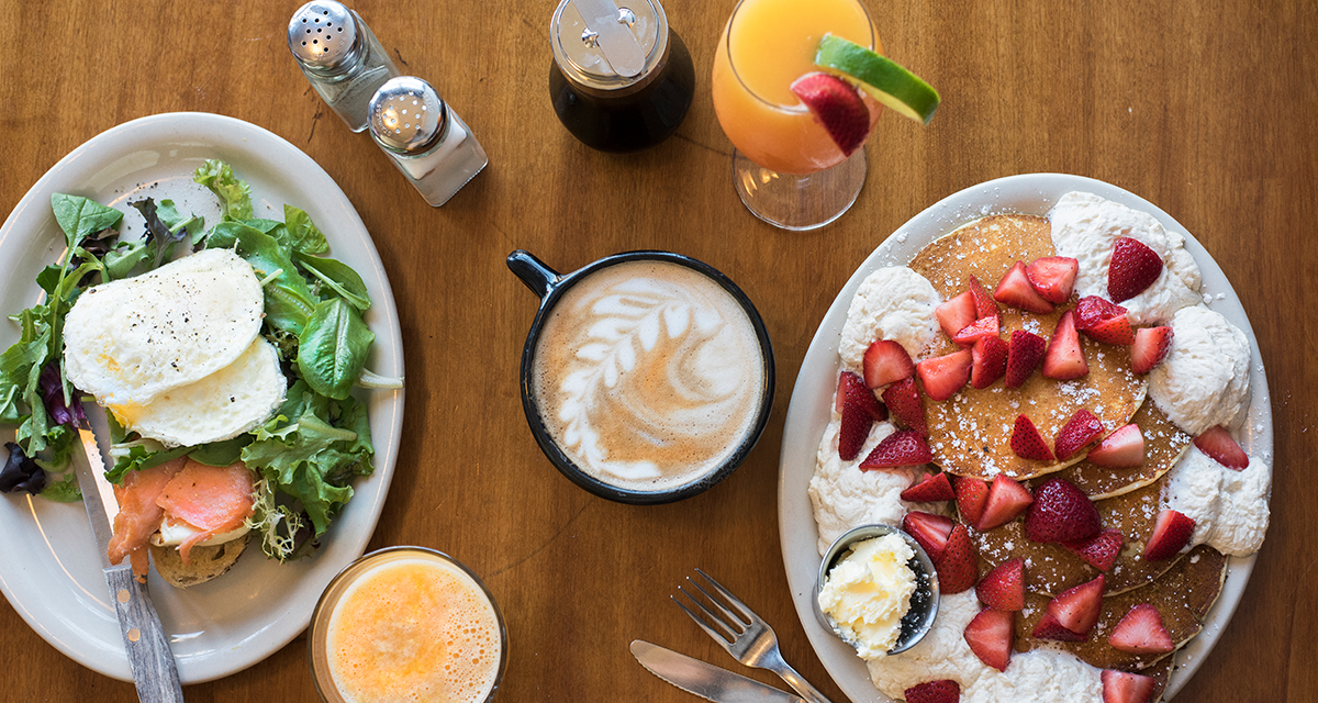 a bird's eye view of a breakfast spread consisting of pancakes with strawberries and whipped cream, eggs, greens, latte art, a mimosa with strawberry and lime, orange juice, syrup dispenser, salt and pepper, and silverware 