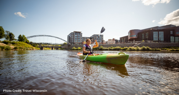 A kayaker floating down the Chippewa River with Haymarket Plaza and Pablo Center in the background 