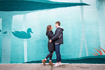A man and woman hugging each other and smiling in front of a big art mural while on a date for Valentine's day in Eau Claire 