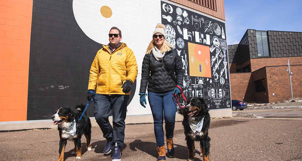 a man and woman couple walking their two dogs in front of the Bon Iver murals in Eau Claire 